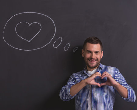 Handsome young guy is showing heart sign, looking at camera and smiling, standing against blackboard with drawn speech bubble and heart in itの写真素材