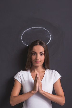 Beautiful young girl is showing angel, holding palms together and looking at camera with tender smile, standing against blackboard with halo overheadの写真素材