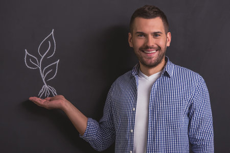 Handsome young man is looking at camera, smiling and pointing at plant drawn on blackboard against...