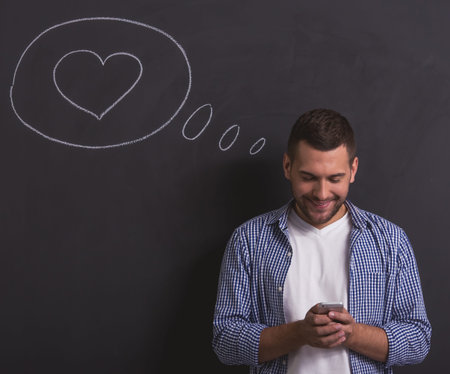 Handsome young guy is texting using a smartphone and smiling, standing against blackboard with drawn speech bubble and heart in itの写真素材