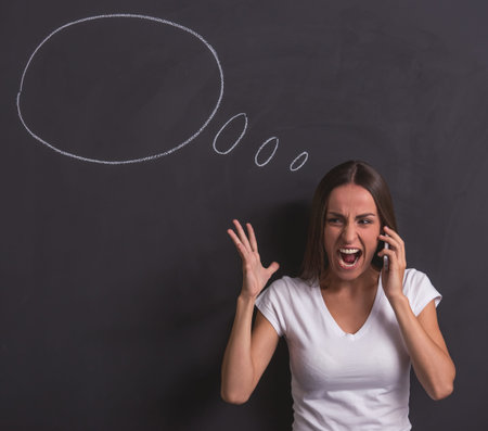 Beautiful young girl is screaming while talking on the mobile phone and gesturing, standing against...
