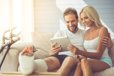 Handsome man with broken leg and his beautiful girlfriend are using a digital tablet and smiling while sitting on couch at homeの写真素材