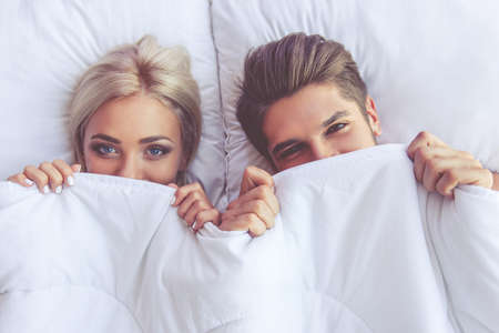Top view of beautiful young couple hiding under the blanket and looking at camera while lying in bedの写真素材