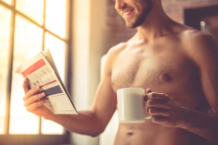 Cropped image of sexy young man with bare torso holding a cup, reading a newspaper and smiling while standing in kitchen at homeの写真素材