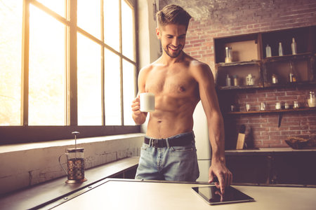 Sexy young man with bare torso is holding a cup, using a digital tablet and smiling while standing in kitchen at homeの写真素材