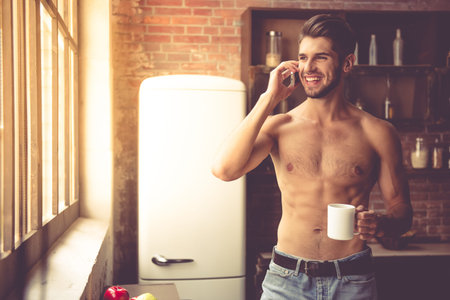 Sexy young man with bare torso is holding a cup, talking on the mobile phone and smiling while standing in kitchen at homeの写真素材