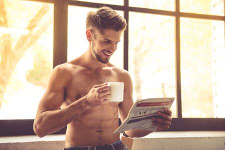 Sexy young man with bare torso is holding a cup, reading a newspaper and smiling while standing in kitchen at homeの写真素材