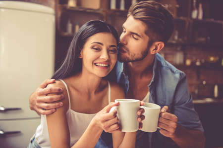 Beautiful young couple is hugging and smiling while drinking tea in kitchen at home. Man is kissing his charming girlの写真素材