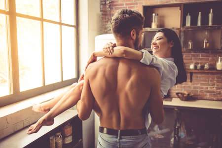 Sexy young man with bare torso is holding his beautiful girlfriend in arms while standing in kitchen at home. Both are looking at each other and smilingの写真素材