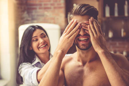 Beautiful young woman is covering her handsome guy eyes. Both are  smiling while standing in kitchen at homeの写真素材
