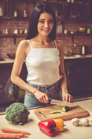 Beautiful young girl in casual clothes is cutting vegetables, looking at camera and smiling while cooking in kitchen at homeの写真素材