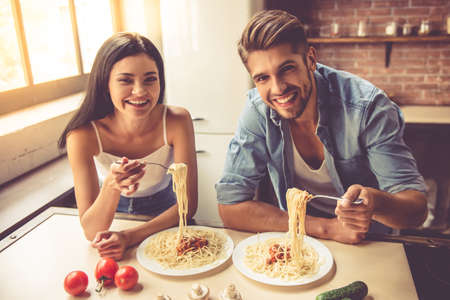 Beautiful young couple is talking, looking at camera and smiling while eating in kitchen at homeの写真素材