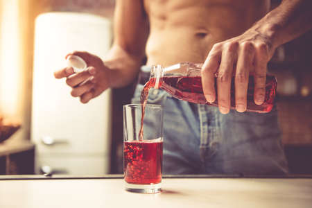 Cropped image of sexy young man with bare torso pouring juice into a glass while standing in kitchen at homeの写真素材