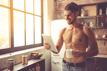 Sexy young man with bare torso is holding a cup, using a digital tablet and smiling while standing in kitchen at homeの写真素材