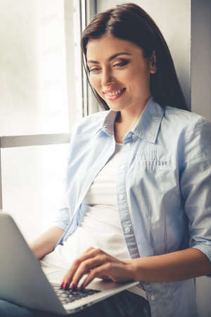 Beautiful girl in casual clothes is using a laptop and smiling while sitting near the window at homeの写真素材