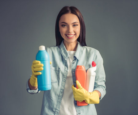 Beautiful young woman in protective gloves is holding detergents, looking at camera and smiling, on gray backgroundの写真素材