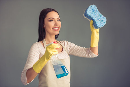 Beautiful young woman is holding a sponge and a spray, looking away and smiling, on gray backgroundの写真素材