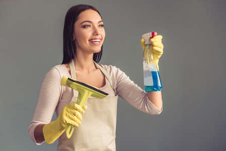Beautiful young woman is holding a squeegee and a spray, looking away and smiling, on gray backgroundの写真素材