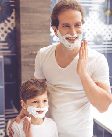 Father and son are applying shaving foam on their faces and smiling while shaving in bathroomの写真素材
