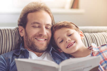 Father and son are reading a book and smiling while spending time together at homeの写真素材