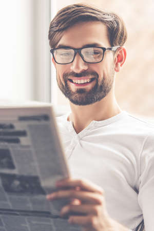 Handsome man in eyeglasses is reading a newspaper and smiling while sitting on couch at homeの写真素材