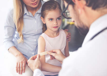 Beautiful young mother and her little daughter at the pediatrician. Doctor is using adhesive bandageの写真素材