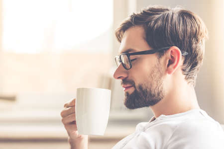 Handsome man in eyeglasses is smelling the aroma of coffee and smiling while resting at homeの写真素材