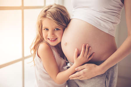 Cute little girl is hugging her beautiful pregnant mom's tummy, looking at camera and smilingの写真素材