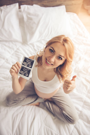 High angle view of beautiful pregnant woman holding a sonogram,showing Ok sign and smiling while...