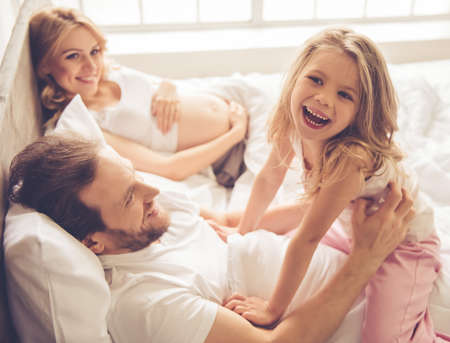 Beautiful pregnant woman, her handsome husband and cute little daughter are smiling while spending time together in bed-roomの写真素材