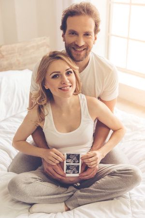 Beautiful pregnant woman and her handsome husband are holding a sonogram, hugging and smiling while sitting on bedの写真素材