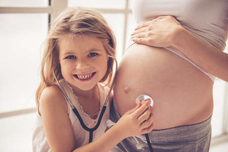 Cute little girl is listening to her beautiful pregnant mom tummy, using a stethoscope, looking at camera and smilingの写真素材