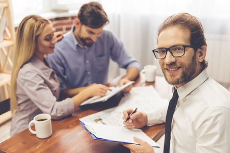Handsome real estate agent in suit is making notes, looking at camera and smiling while happy beautiful couple is talking in the backgroundの写真素材
