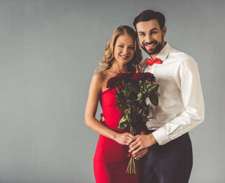 Beautiful elegant girl in red dress and guy in classic shirt with red bow tie are holding roses and smiling, on gray backgroundの写真素材