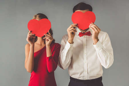 Beautiful elegant girl in red dress and guy in classic shirt and red bow tie are holding paper hearts, on gray backgroundの写真素材