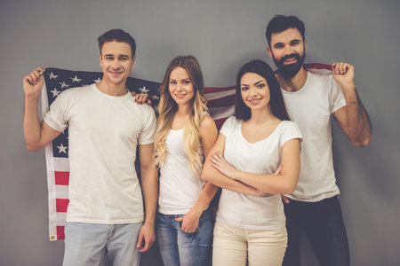 Beautiful young friends are holding an American flag, looking at camera and smiling, on gray...