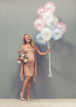 Full length portrait of beautiful pregnant woman holding balloons and teddy bear, looking at camera and smiling, on gray backgroundの写真素材