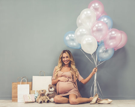 Beautiful pregnant woman holding balloons, looking at camera and smiling while sitting near her...