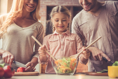 Cute little girl and her beautiful parents are cutting vegetables and smiling while making salad in kitchen at homeの写真素材