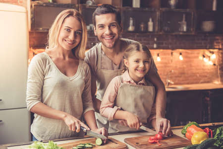 Cute little girl and her beautiful parents are cutting vegetables and smiling while cooking in kitchen at homeの写真素材
