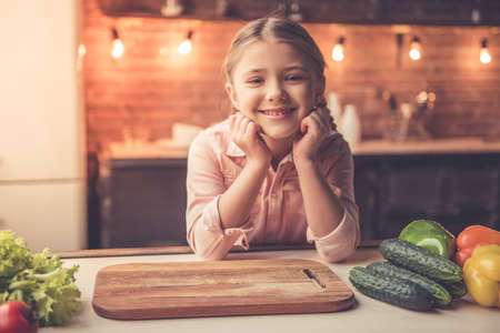 Cute little girl is looking at camera and smiling while cooking in kitchen at homeの写真素材
