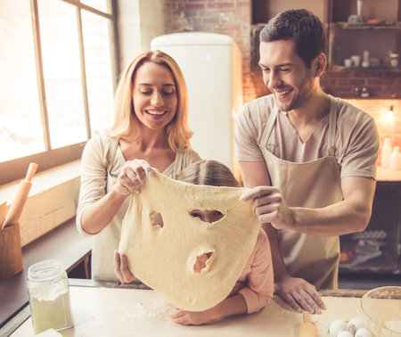 Cute little girl and her beautiful parents in aprons are making dough and smiling while baking in...