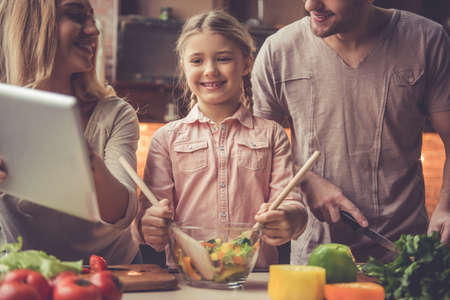 Cute little girl and her beautiful parents are using digital tablet and smiling while cooking in kitchen at homeの写真素材