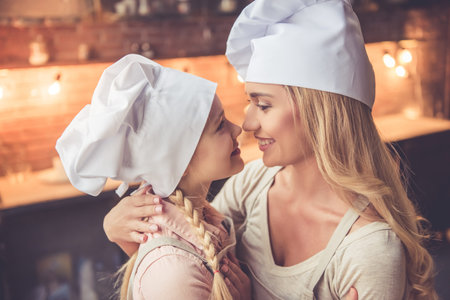 Cute little girl and her beautiful mom in chef's hats are touching noses and smiling while cooking in kitchen at homeの写真素材