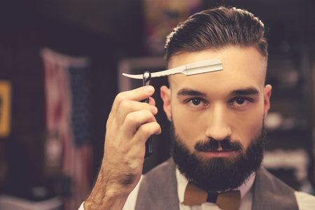 Handsome bearded hairdresser in stylish classic wear is holding a straight razor while standing at...