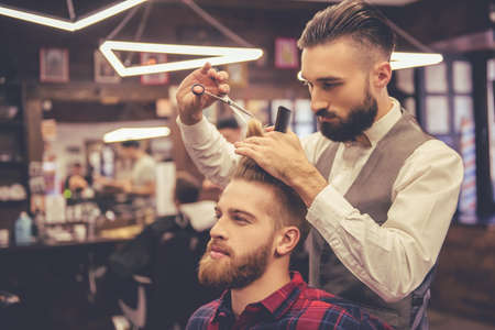 Handsome bearded man is looking forward while having his hair cut by hairdresser at the barbershopの写真素材