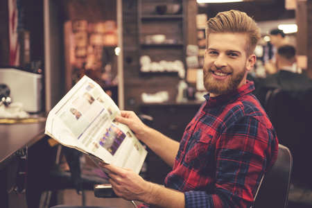 Handsome bearded man is reading a newspaper, looking at camera and smiling while sitting in chair at the barbershopの写真素材