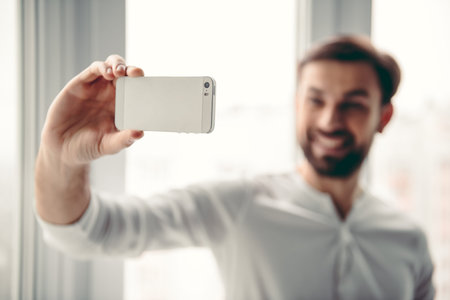 Handsome young man is doing selfie using a smart phone and smiling while standing near the window....