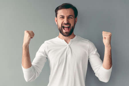 Handsome young man is raising hands in fists and screaming with happiness, on gray backgroundの写真素材
