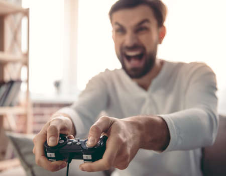 Handsome young man is playing game console while sitting on couch at homeの写真素材